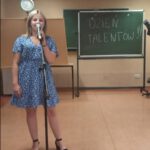 Dziewczynka w niebiesko-białej sukience stoi przy mikrofonie i śpiewa. Za nią stoi tablica z napisem : Dzień Talentów"