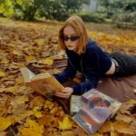 Dziewczyna w ciemnych okularach leży na kocu w jesiennej scenerii i czyta książkę